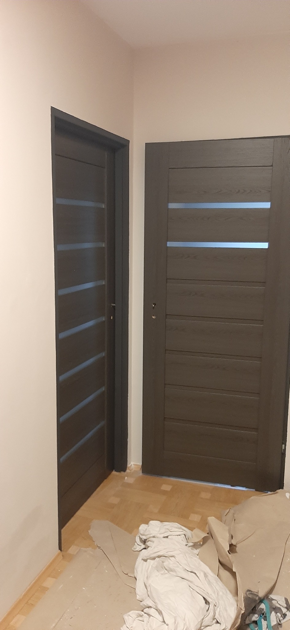 Dwa ciemne, drewniane drzwi wewnętrzne z poziomymi, przeszklonymi paskami, widoczne po remoncie, z zabezpieczeniami na podłodze.