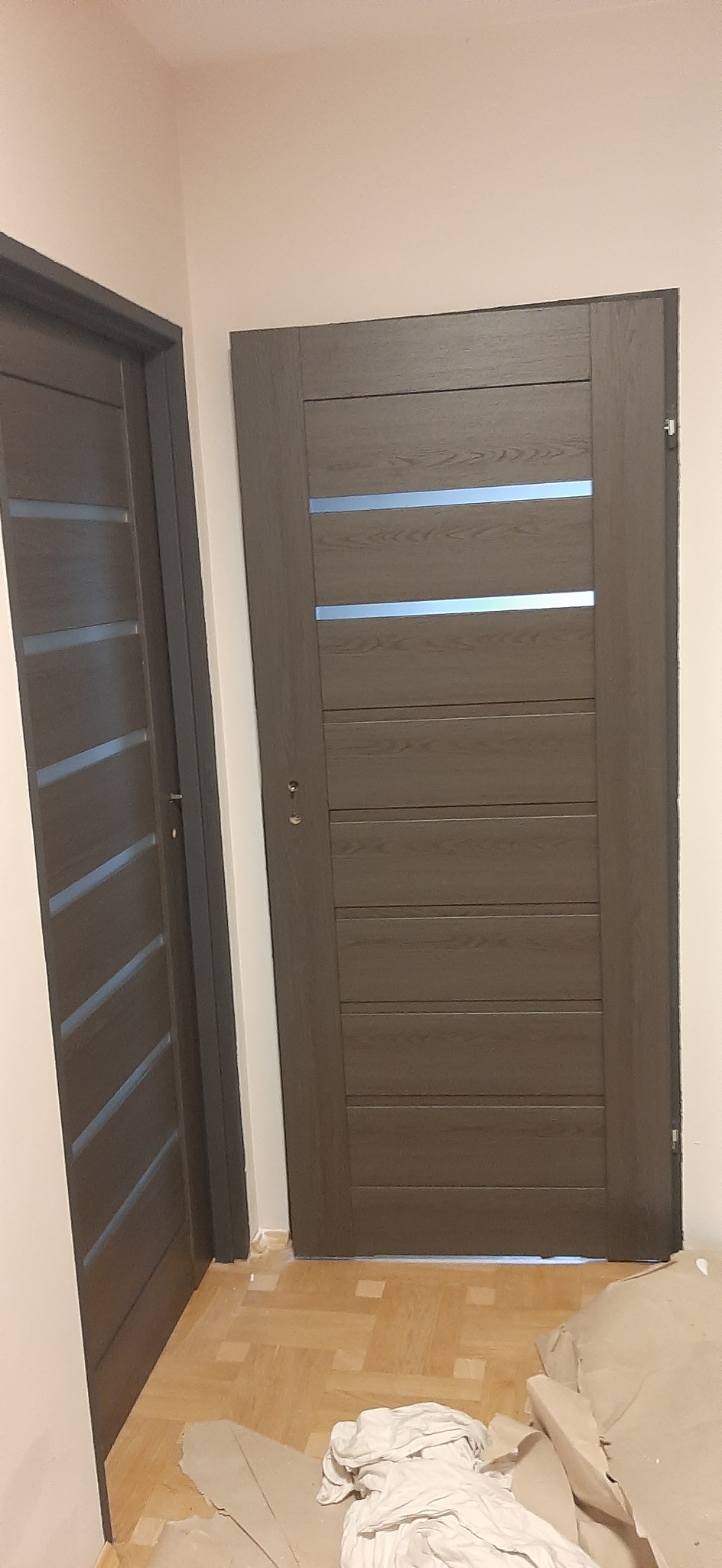 Dwa brązowe, drewniane drzwi wewnętrzne z poziomymi, szklanymi wstawkami, świeżo zamontowane na parkiecie. Widoczne ślady po remoncie.