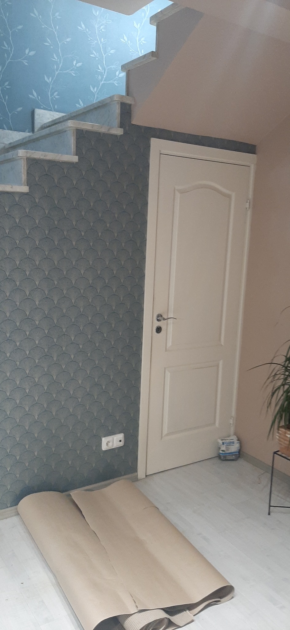 Szary wzorzysty panel ścienny z niebieskimi akcentami pod schodami, białe stopnie, białe drzwi, zwinięta wykładzina podłogowa i paczka gipsu