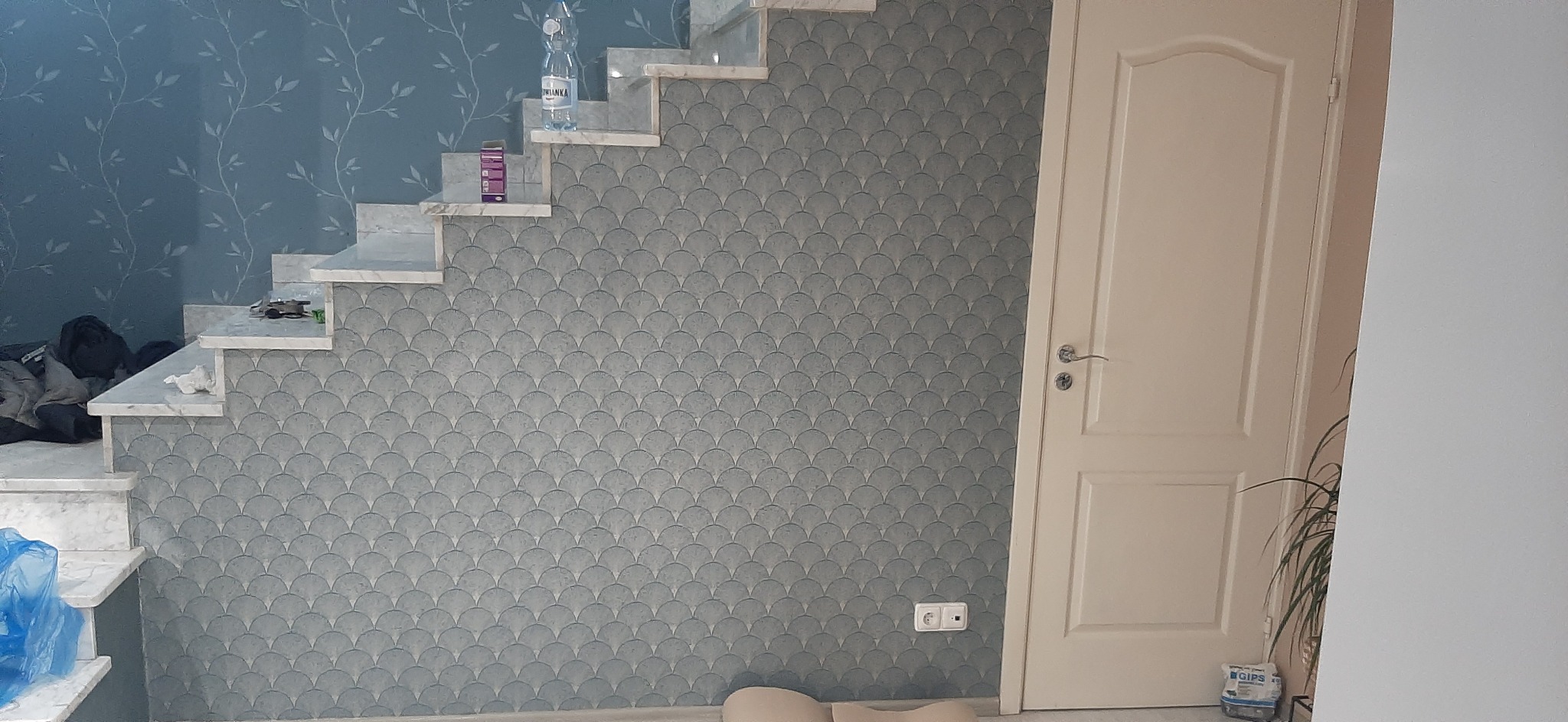 Ściana z dekoracyjną tapetą w łuski obok marmurowych schodów i białych drzwi z widocznym gniazdkiem elektrycznym.