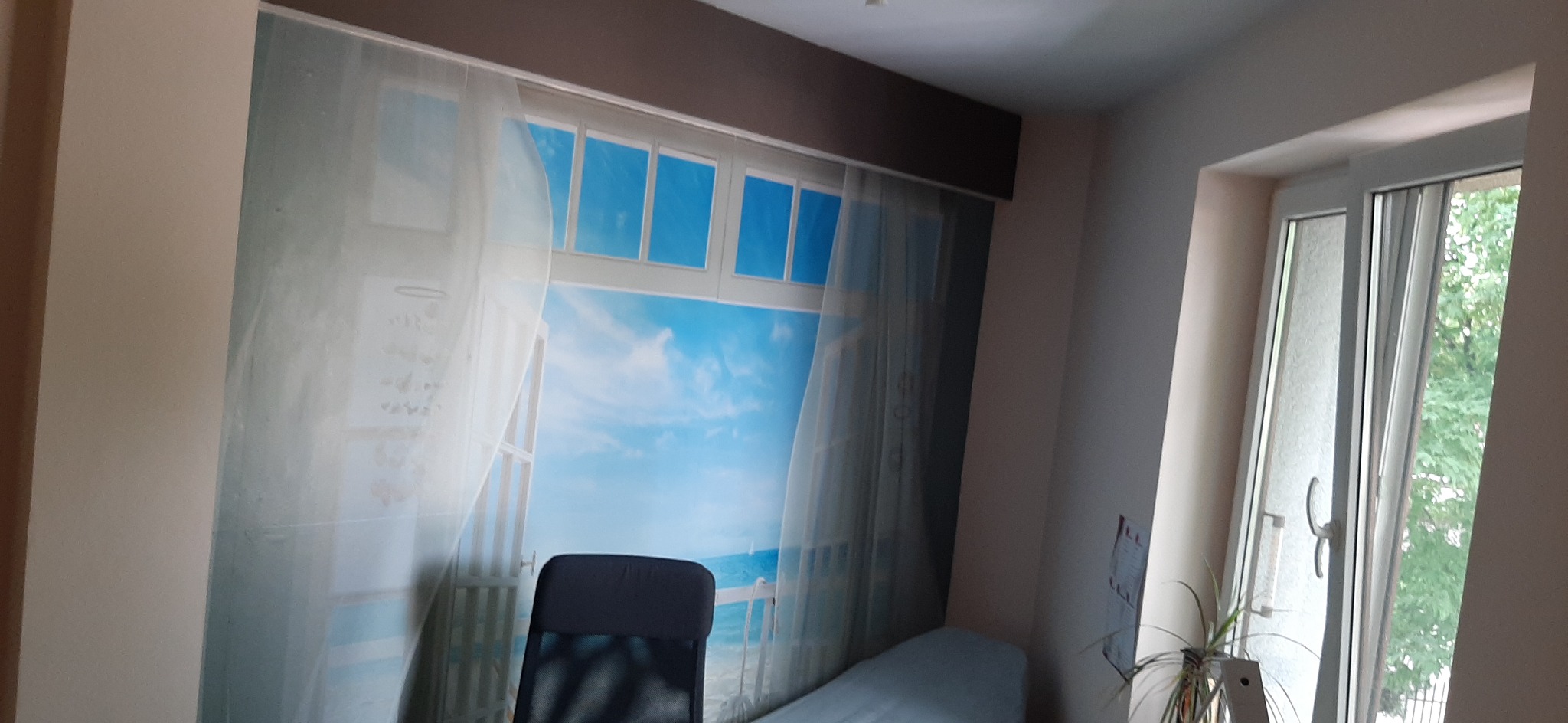 Fototapeta z motywem okna z widokiem na morze, naklejona na ścianę w pokoju, z widocznym fragmentem łóżka i krzesła na pierwszym planie.