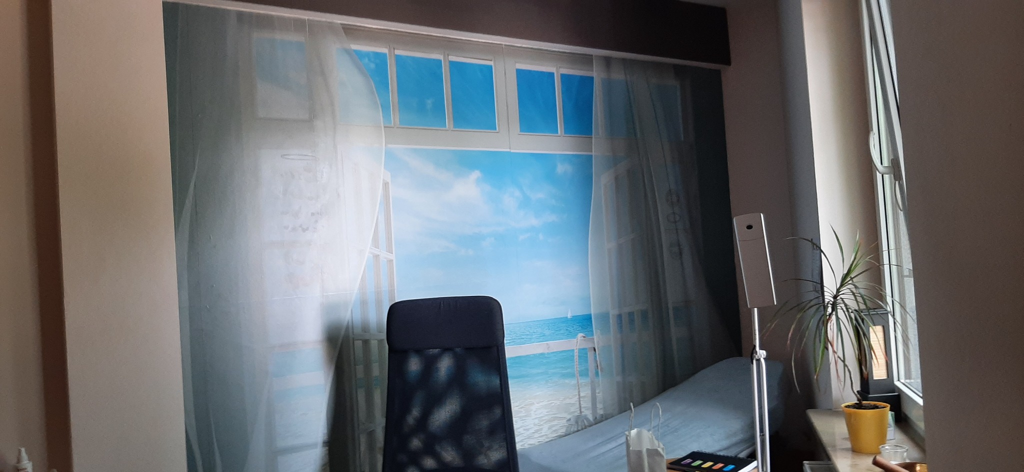 Fototapeta z motywem okna z widokiem na plażę i morze, zamontowana na ścianie w pokoju, z widocznym biurkiem i rośliną doniczkową.