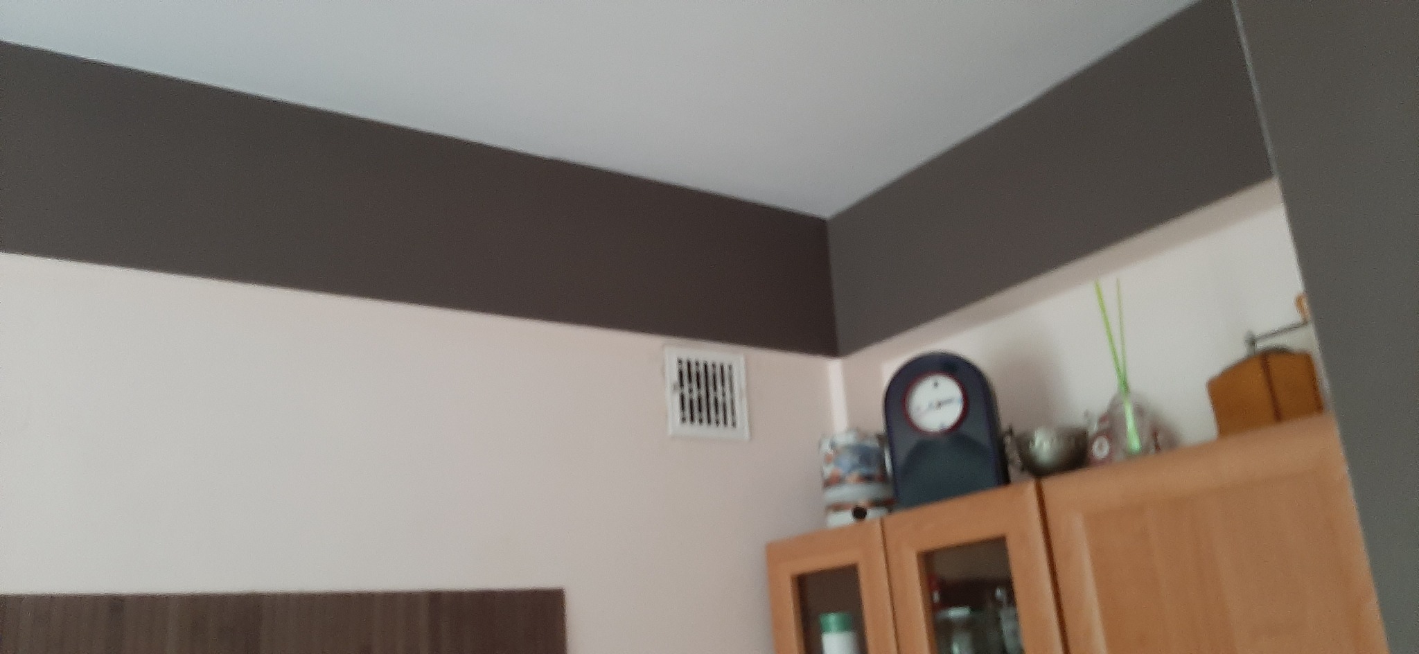 Pomieszczenie po odświeżeniu: ściany pomalowane na jasny róż, z szerokim, poziomym pasem w kolorze ciemnego brązu pod sufitem, widoczna kratka wentylacyjna i szafki kuchenne.