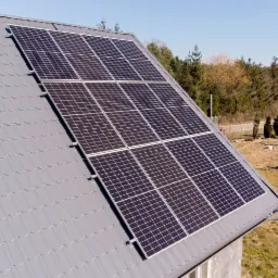 Panele słoneczne zamontowane na szarym dachu z blachodachówki, widok z góry na tle drzew i błękitnego nieba.