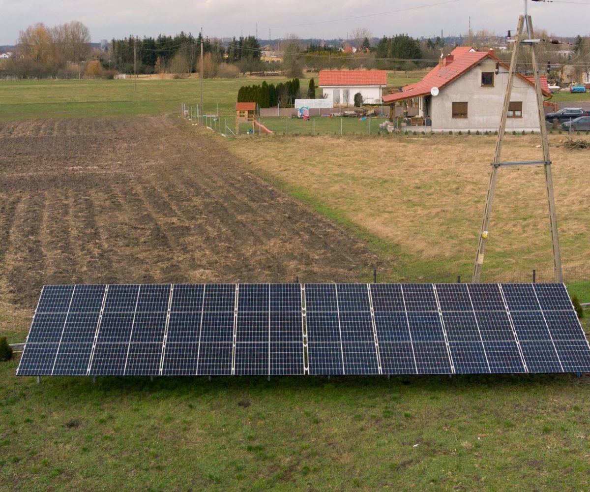 Instalacja paneli słonecznych na gruncie przed domem jednorodzinnym z czerwonym dachem, widok z lekkiej góry na panele ustawione rzędami na zielonej trawie, w tle pole i słup energetyczny.