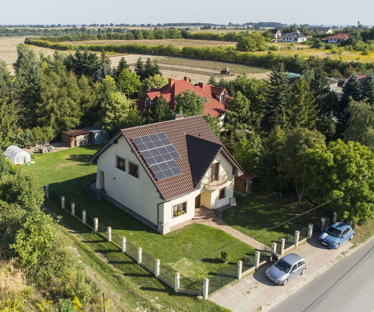 Dom jednorodzinny z brązowym dachem i panelami słonecznymi, widok z lotu ptaka, otoczony zielenią i dwoma samochodami na podjeździe.