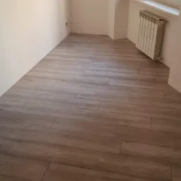 Ułożone panele podłogowe imitujące drewno w pustym pokoju w Przecławiu, widoczny grzejnik pod oknem i wystające kable ze ściany.