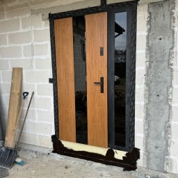 VIRTUS SYSTEM - Montaż drzwi zewnętrznych z drewnopodobnym wykończeniem, widoczne narzędzia i uszczelnianie pianką montażową, surowy stan budynku w tle.