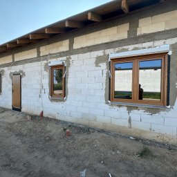 VIRTUS SYSTEM - Surowy stan budynku w trakcie montażu stolarki okiennej i drzwiowej: brązowe ramy okienne z zamontowanymi roletami zewnętrznymi, brązowe drzwi wejściowe, ściany z białych bloczków, ziemia wokół.