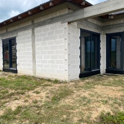 VIRTUS SYSTEM - Surowy stan budynku z szarego betonu komórkowego z zamontowanymi czarnymi oknami PCV, widok z zewnątrz na trawiasty teren.