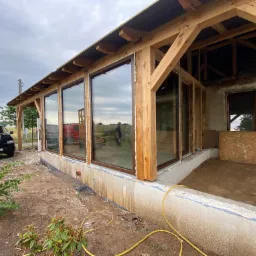 Montaż dużych okien w drewnianej konstrukcji budynku w trakcie budowy, widoczne pianowanie i surowy stan wykończenia.