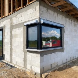 VIRTUS SYSTEM - Surowy budynek z białych bloczków, z zamontowanymi oknami z czarnymi ramami i szarymi roletami zewnętrznymi, widoczne rusztowanie dachu, teren wokół w trakcie prac ziemnych.