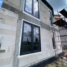 VIRTUS SYSTEM - Montaż czarnych ram okiennych w surowym budynku z białych bloczków w Biskupcu, widoczne rusztowanie i odbicie drzewa w szybach.