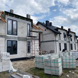 VIRTUS SYSTEM - Budowa bliźniaka z zamontowanymi oknami w czarnych ramach, widoczne rusztowania, palety z bloczkami betonowymi i czerwony samochód dostawczy w tle.