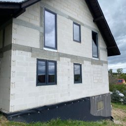 VIRTUS SYSTEM - Surowy stan domu w trakcie budowy, widoczne wstawione grafitowe ramy okienne w ścianie z białych bloczków, czarna folia fundamentowa, pochmurne niebo.