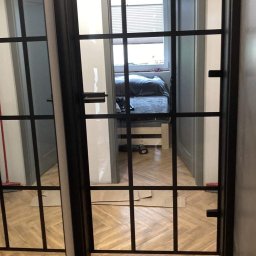 VIRTUS SYSTEM - Czarne, przeszklone drzwi wewnętrzne z kratownicą, widok na sypialnię z parkietem w jodełkę, częściowo widoczne narzędzia i materiały montażowe na podłodze.