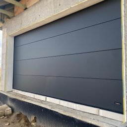 VIRTUS SYSTEM - Szara segmentowa brama garażowa z poziomymi przetłoczeniami, zamontowana w surowym otworze budynku w trakcie budowy, widoczne bloczki betonowe i izolacja, w tle pole i turbina wiatrowa.