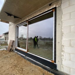 VIRTUS SYSTEM - Montaż dużego okna przesuwnego z brązową ramą w surowym stanie budynku, widoczne narzędzia i odbicie dwóch osób w szybie.