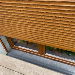 Brązowa roleta zwinięta częściowo na oknie z drewnianą ramą, widoczny fragment rusztowania i surowy tynk na ścianie.