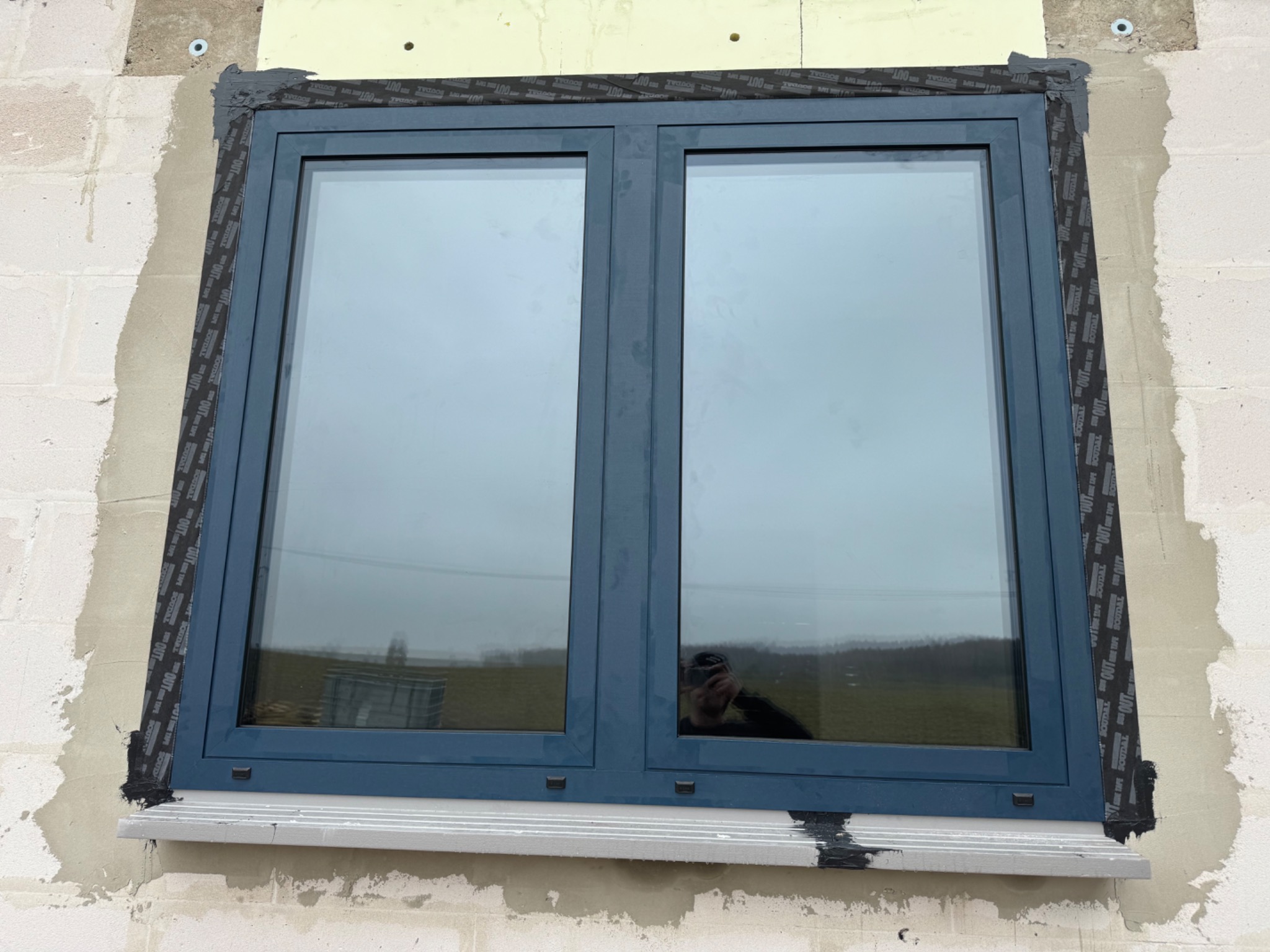 Szare, dwuskrzydłowe okno zamontowane w surowej ścianie z betonu komórkowego, zabezpieczone czarną taśmą uszczelniającą i częściowo obrobione tynkiem. W szybie okna widoczne odbicie fotografa.