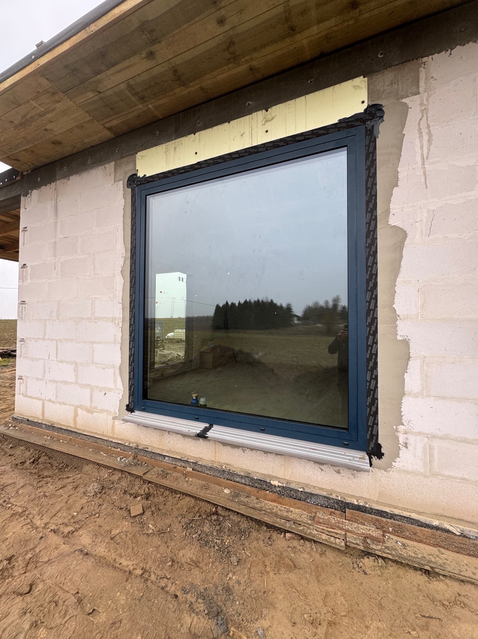 Niebieska rama okienna w surowym stanie, widoczne pianka izolacyjna i taśma uszczelniająca na tle ściany z białych bloczków i grząskiego terenu.