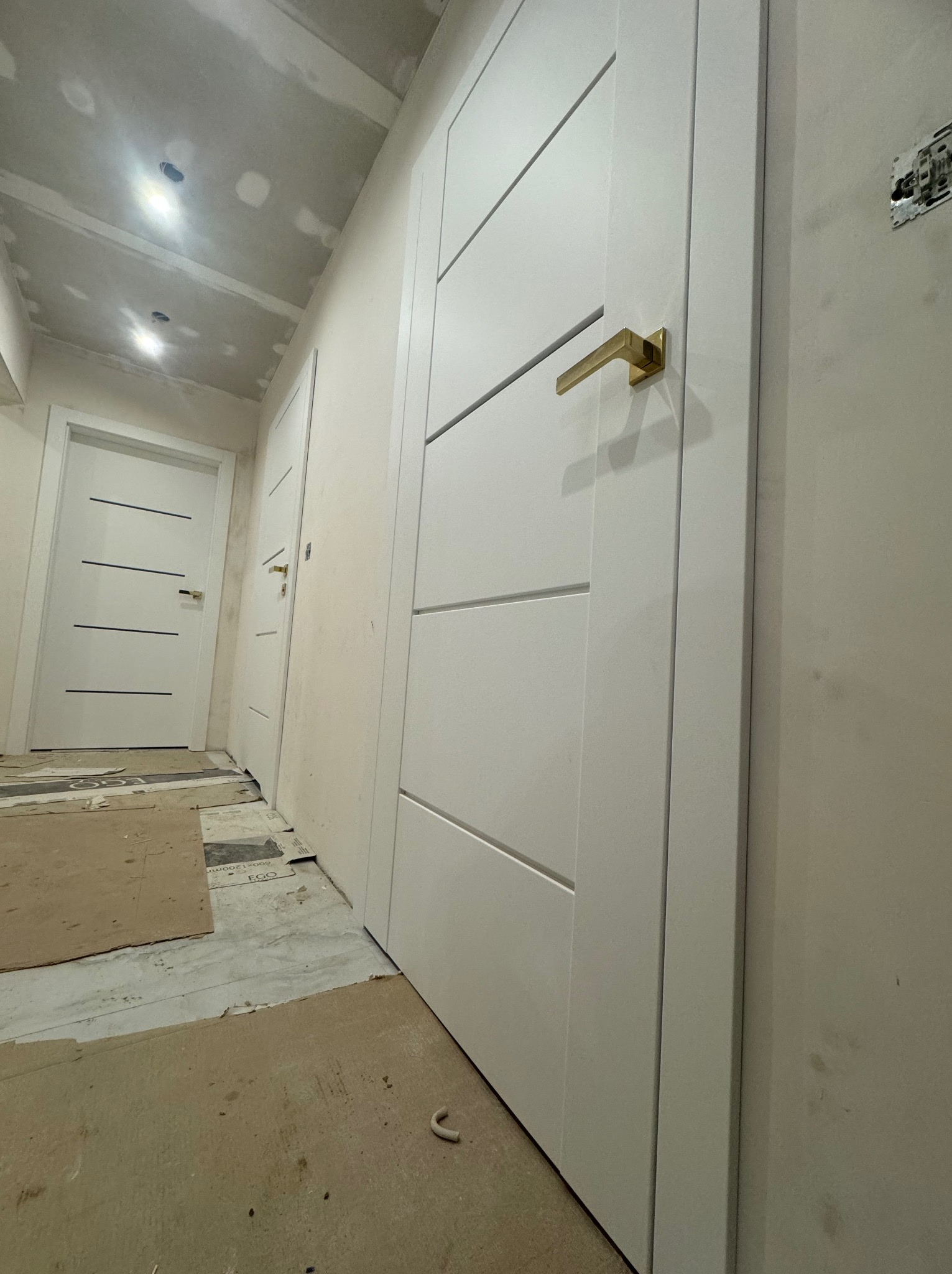 Białe drzwi wewnętrzne z poziomymi frezowaniami i złotą klamką w surowym wnętrzu w trakcie wykończenia, widoczne kartony zabezpieczające podłogę i ślady po gładzi na ścianach.