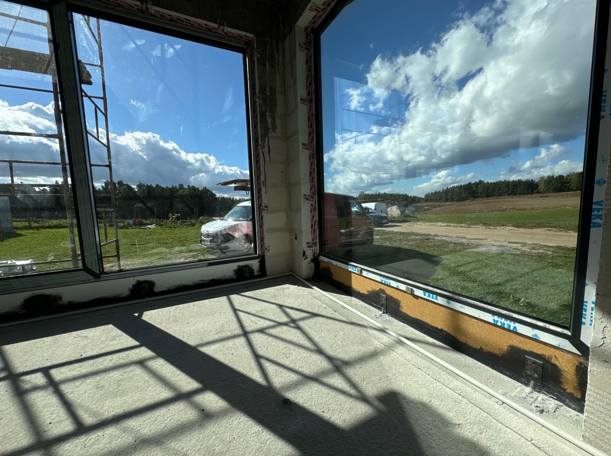 Surowy betonowy pokój z dwoma dużymi oknami z widokiem na zielony krajobraz i błękitne niebo z chmurami; widoczne ślady montażu okien.