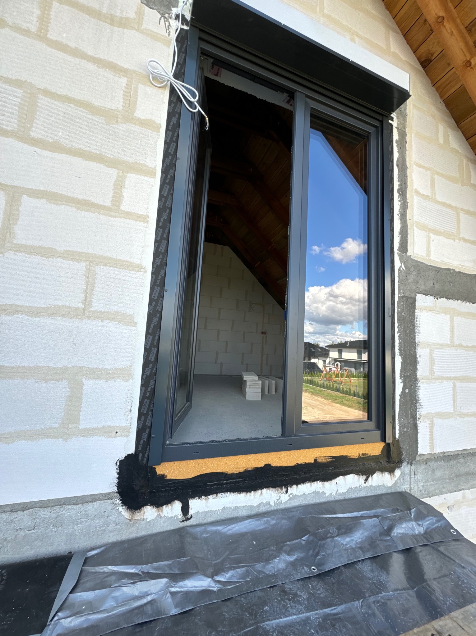 Montaż okna w surowym stanie, widoczne bloczki z betonu komórkowego, czarna izolacja wokół ramy okiennej, częściowo opuszczona roleta zewnętrzna, odbicie nieba z chmurami w szybie okna.