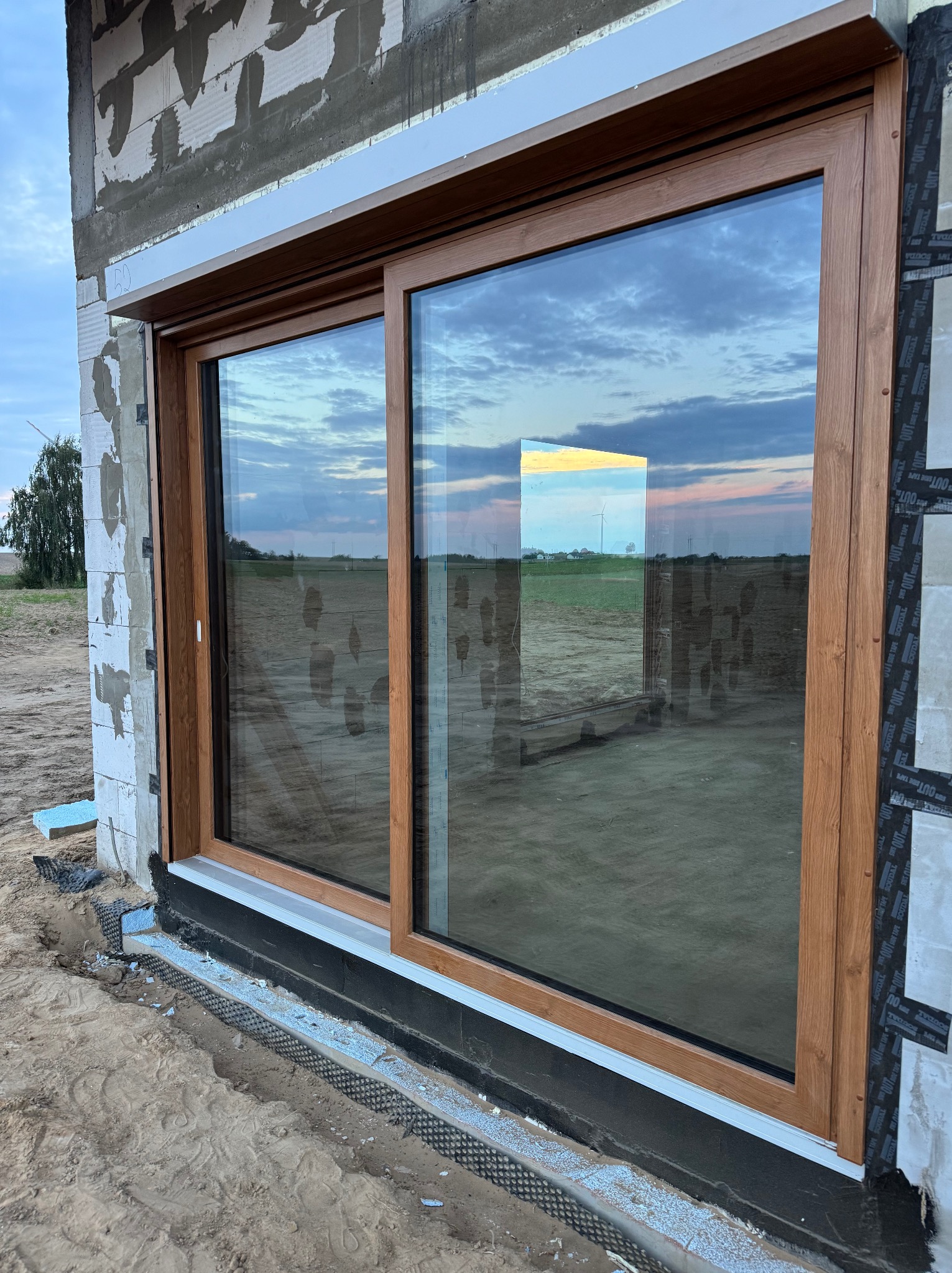 Montaż okna przesuwnego z brązową ramą na surowym murze, widoczne detale izolacji i odblask zachodzącego słońca w szybie.