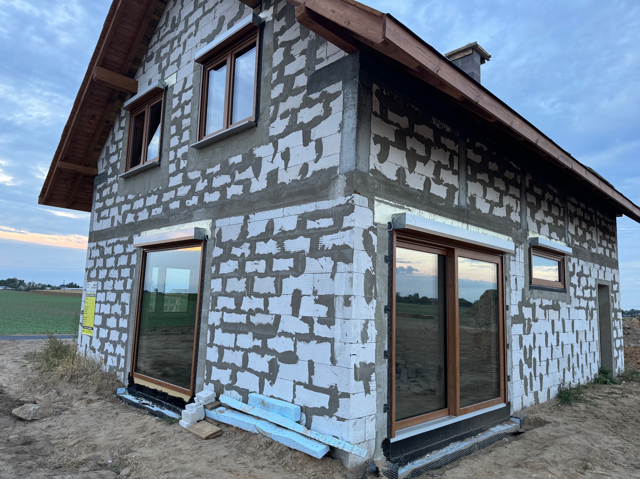 Surowy stan domu w trakcie budowy: widoczne okna PCV z brązowymi ramami zamontowane w ścianach z betonu komórkowego, ocieplenie styropianem w dolnej części, drewniana konstrukcja dachu, teren...