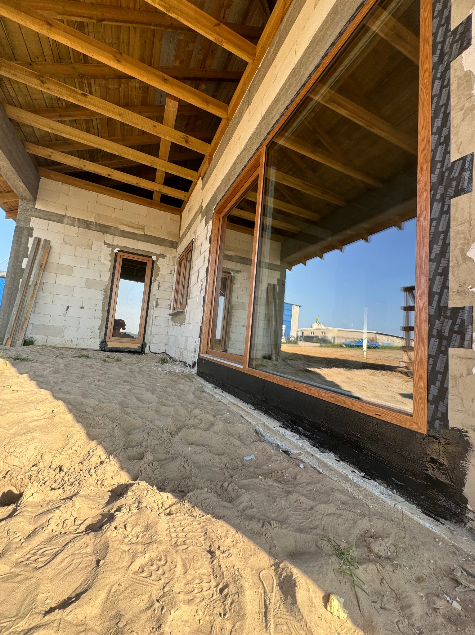 Montaż stolarki okiennej z drewnianą ramą w surowym stanie budynku w Biskupcu, widoczne zabezpieczenie przeciwwilgociowe i piasek wokół budynku.