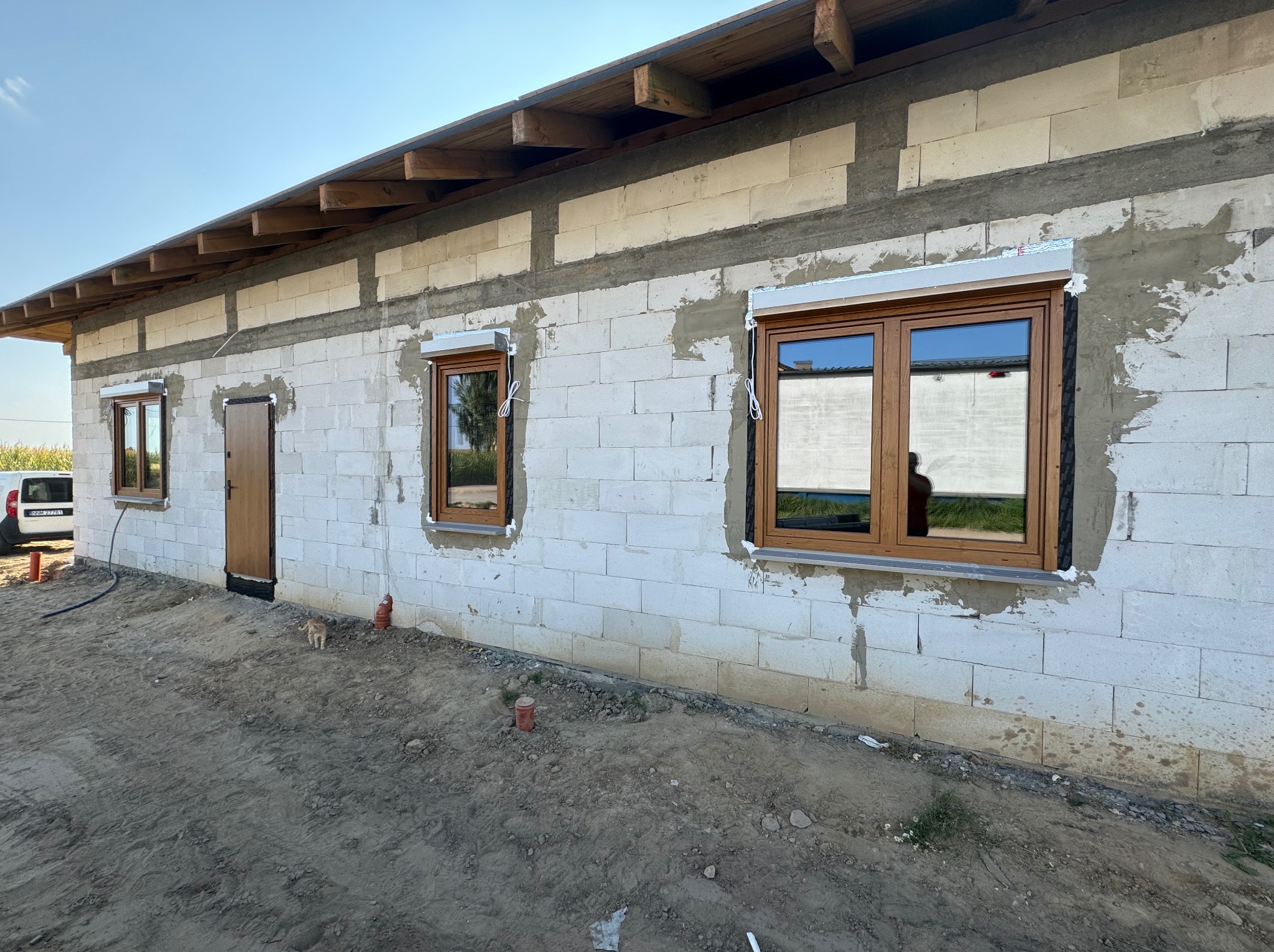 Surowy stan budynku w trakcie montażu stolarki okiennej i drzwiowej: brązowe ramy okienne z zamontowanymi roletami zewnętrznymi, brązowe drzwi wejściowe, ściany z białych bloczków, ziemia wokół.
