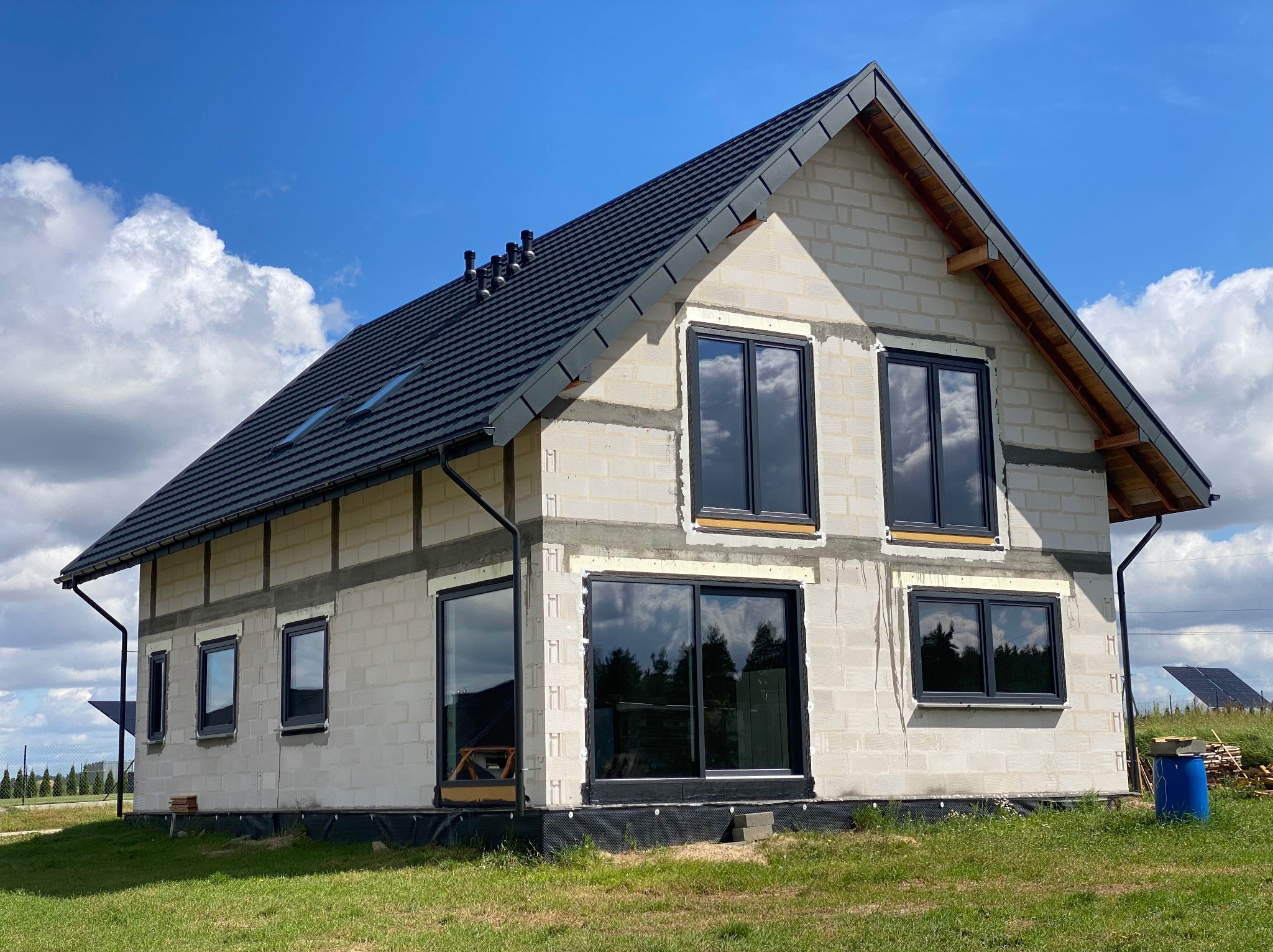 Dwukondygnacyjny dom w trakcie budowy z zamontowanymi oknami w czarnych ramach, widoczne bloczki betonowe, czarny dach z dachówkami i oknami dachowymi, trawa i błękitne niebo w tle.