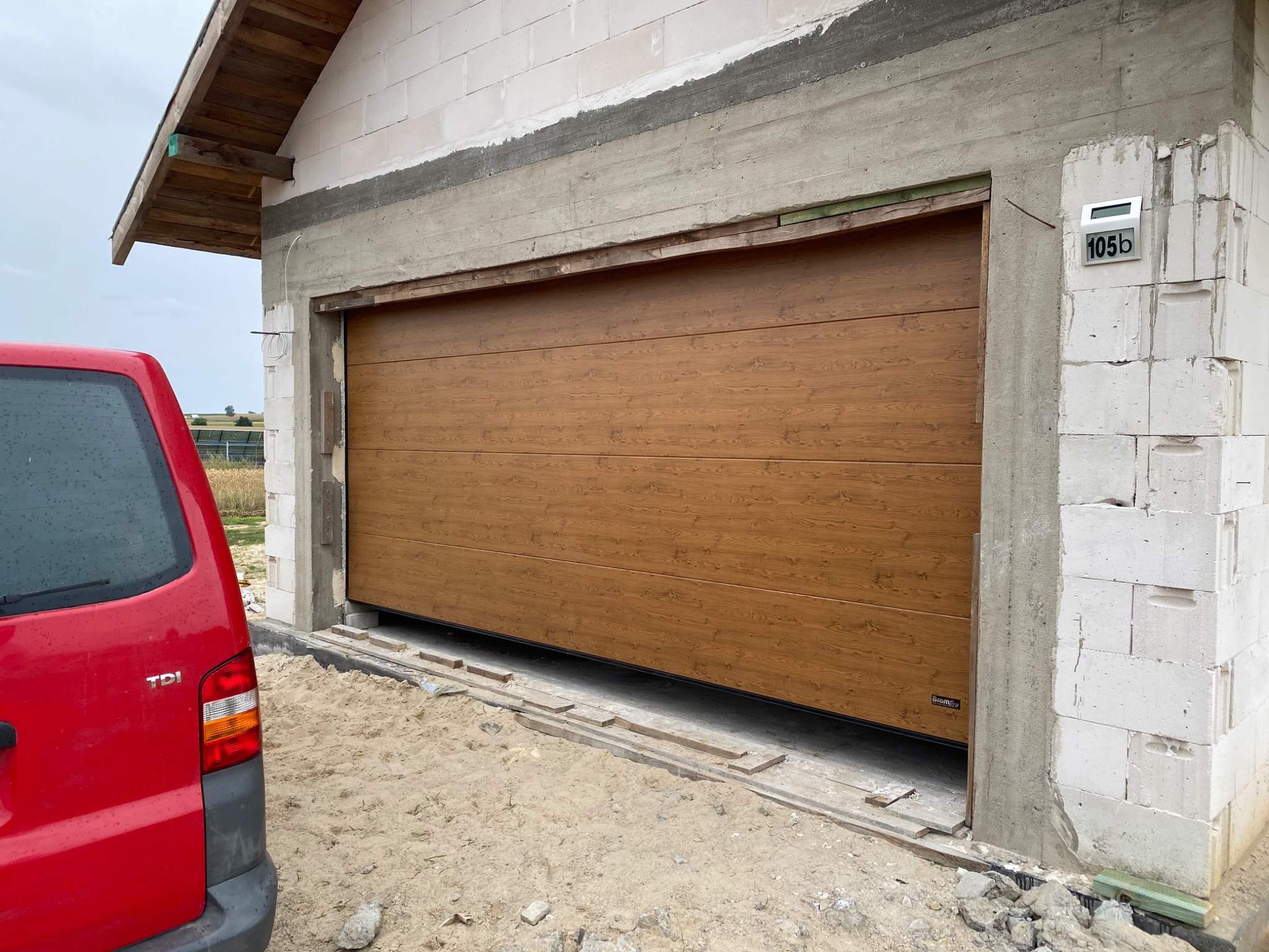 Montaż segmentowej bramy garażowej o wzorze drewna w surowym otworze budynku z czerwonym samochodem dostawczym w tle.