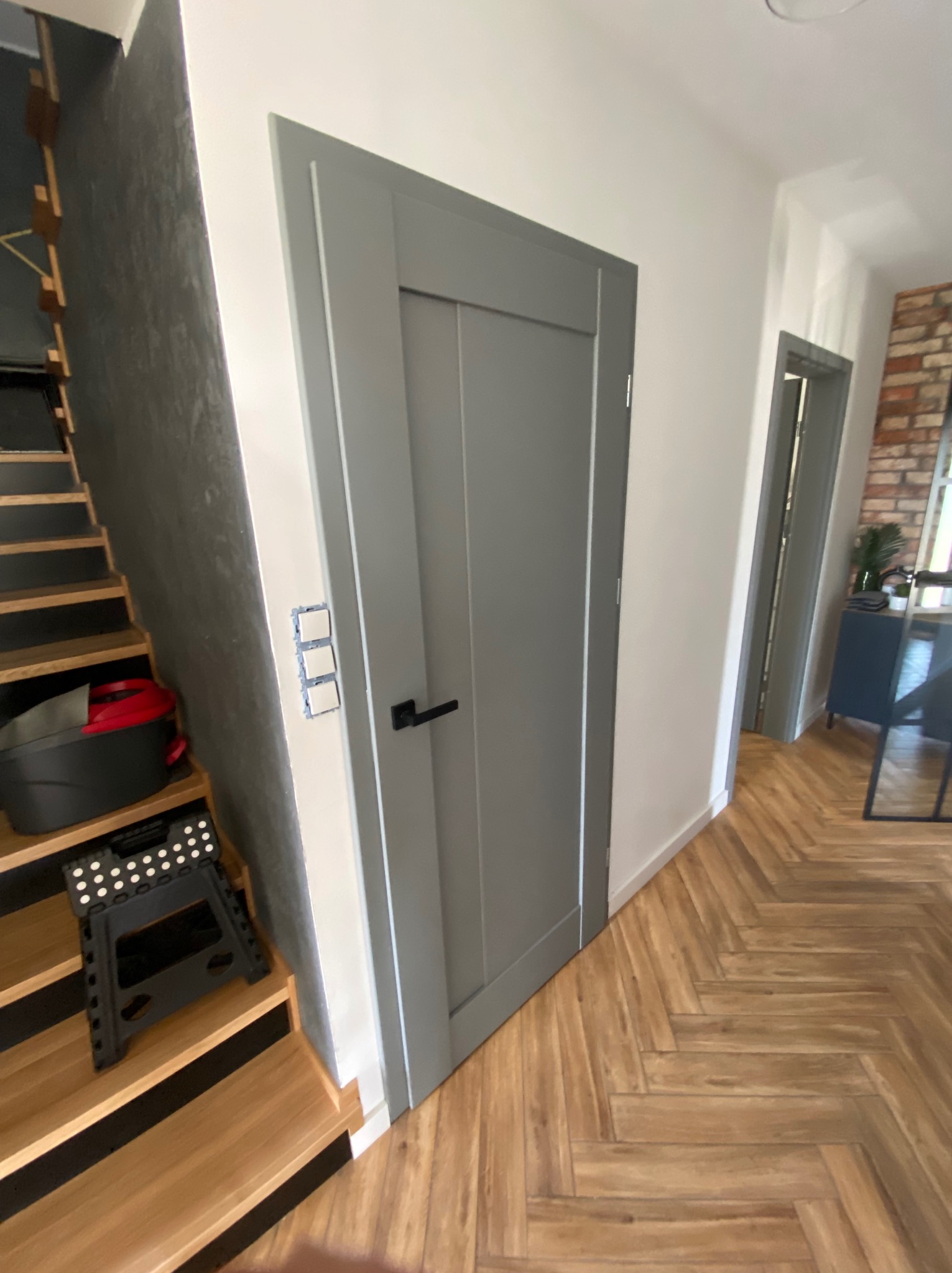 Wnętrze z szarą, panelową drzwią wewnętrzną z czarną klamką, widok na schody z wiaderkiem i taboretem oraz parkiet ułożony w jodełkę.