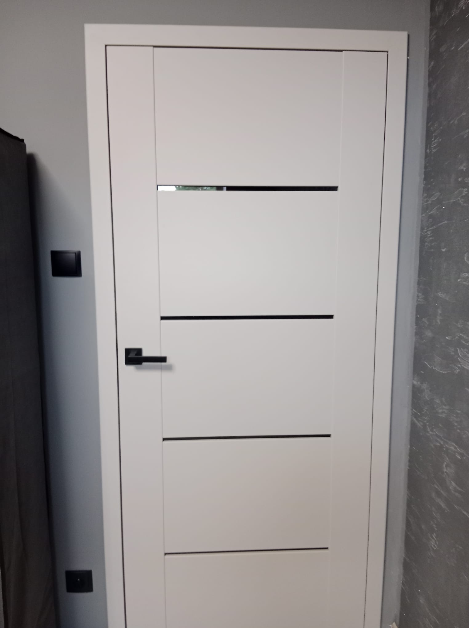 Białe drzwi wewnętrzne z czarną klamką i poziomymi wstawkami na tle szarej ściany z widocznym włącznikiem światła.