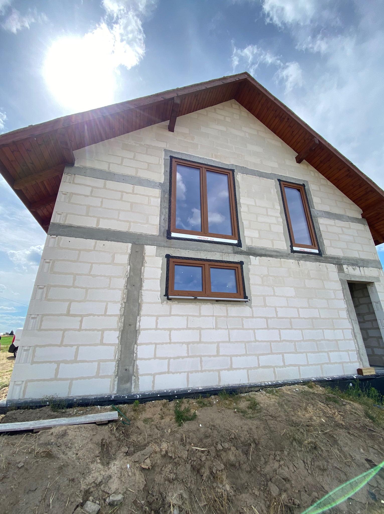 Surowy stan budynku z białych bloczków, widoczne drewniane elementy dachu i brązowe ramy okienne, częściowo osadzone w ścianie, widok z dołu na tle błękitnego nieba z chmurami.