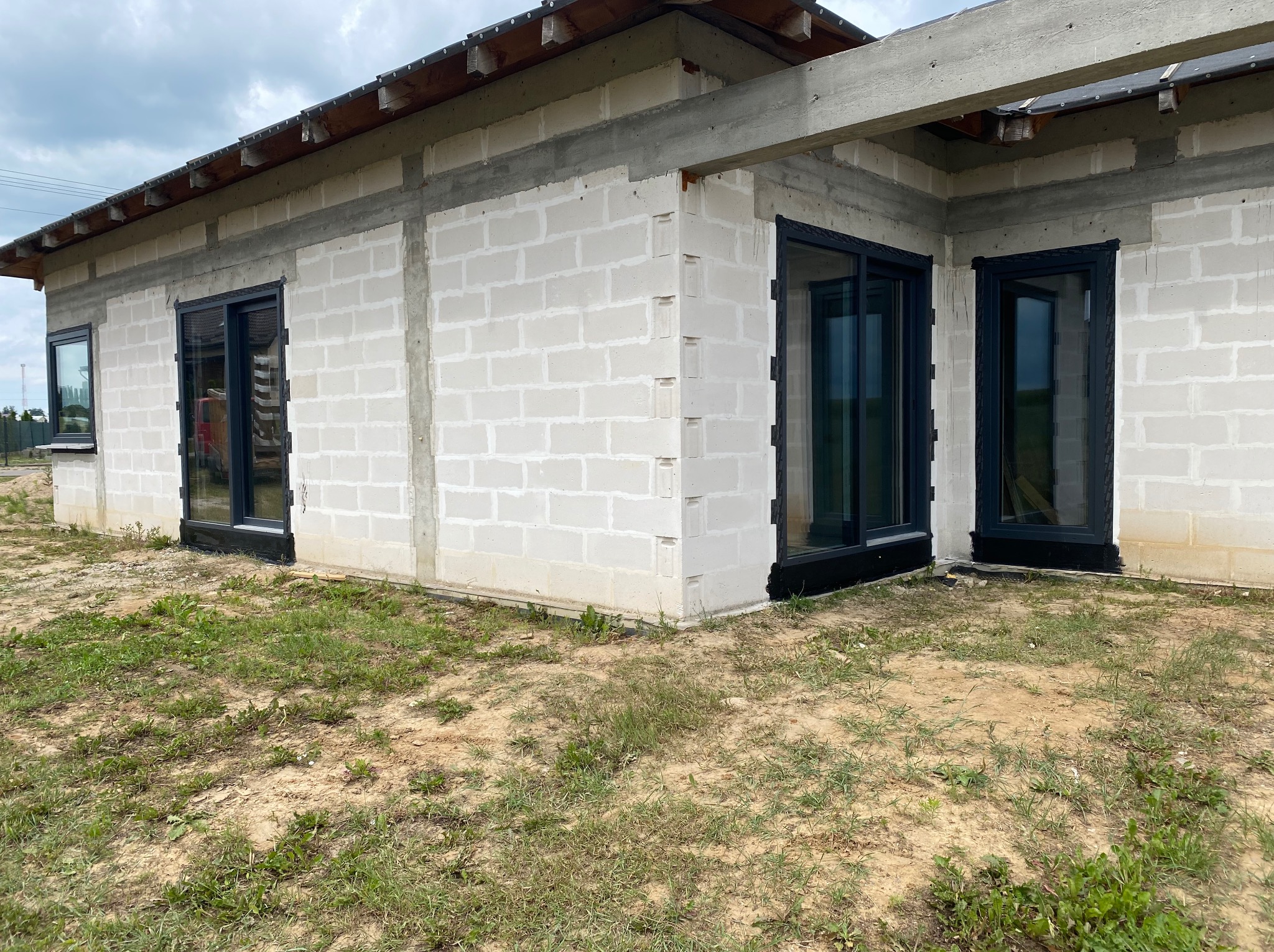 Surowy stan budynku z szarego betonu komórkowego z zamontowanymi czarnymi oknami PCV, widok z zewnątrz na trawiasty teren.