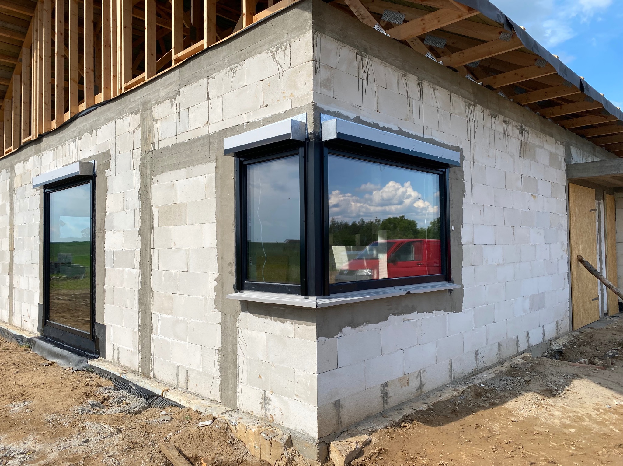 Surowy budynek z białych bloczków, z zamontowanymi oknami z czarnymi ramami i szarymi roletami zewnętrznymi, widoczne rusztowanie dachu, teren wokół w trakcie prac ziemnych.
