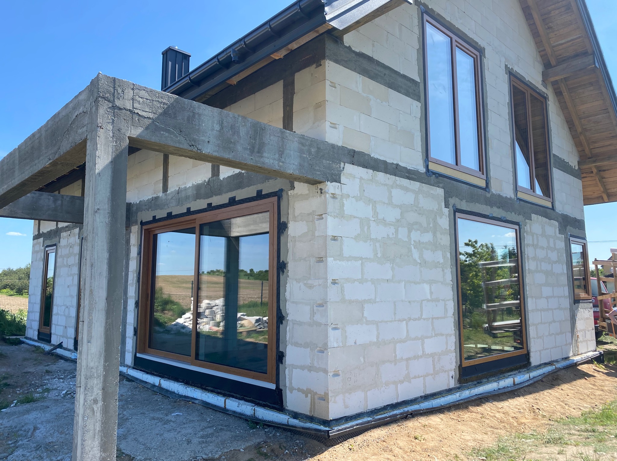 Surowy stan budynku w trakcie budowy z zamontowanymi oknami z brązowymi ramami, widoczne bloczki betonowe i surowy beton pergoli, błękitne niebo.