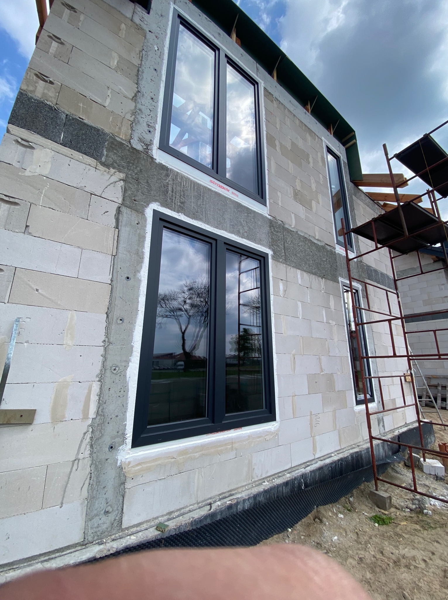 Montaż czarnych ram okiennych w surowym budynku z białych bloczków w Biskupcu, widoczne rusztowanie i odbicie drzewa w szybach.