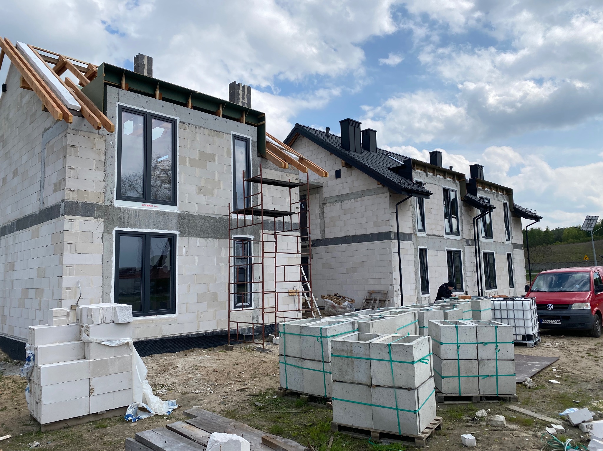 Budowa bliźniaka z zamontowanymi oknami w czarnych ramach, widoczne rusztowania, palety z bloczkami betonowymi i czerwony samochód dostawczy w tle.
