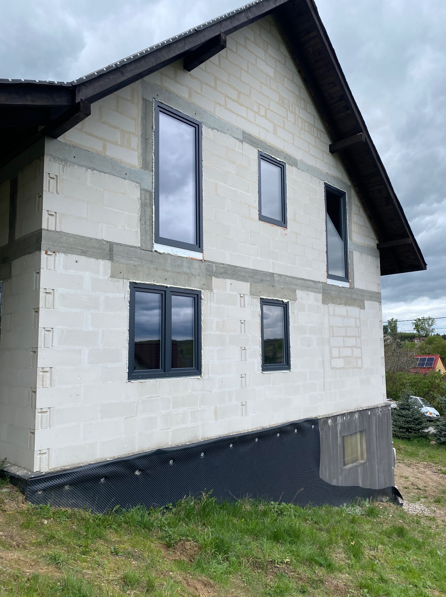 Surowy stan domu w trakcie budowy, widoczne wstawione grafitowe ramy okienne w ścianie z białych bloczków, czarna folia fundamentowa, pochmurne niebo.