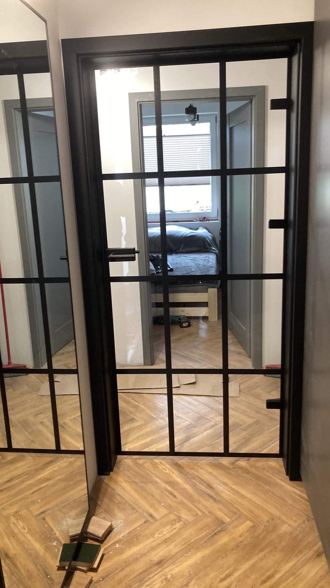 Czarne, przeszklone drzwi wewnętrzne z kratownicą, widok na sypialnię z parkietem w jodełkę, częściowo widoczne narzędzia i materiały montażowe na podłodze.