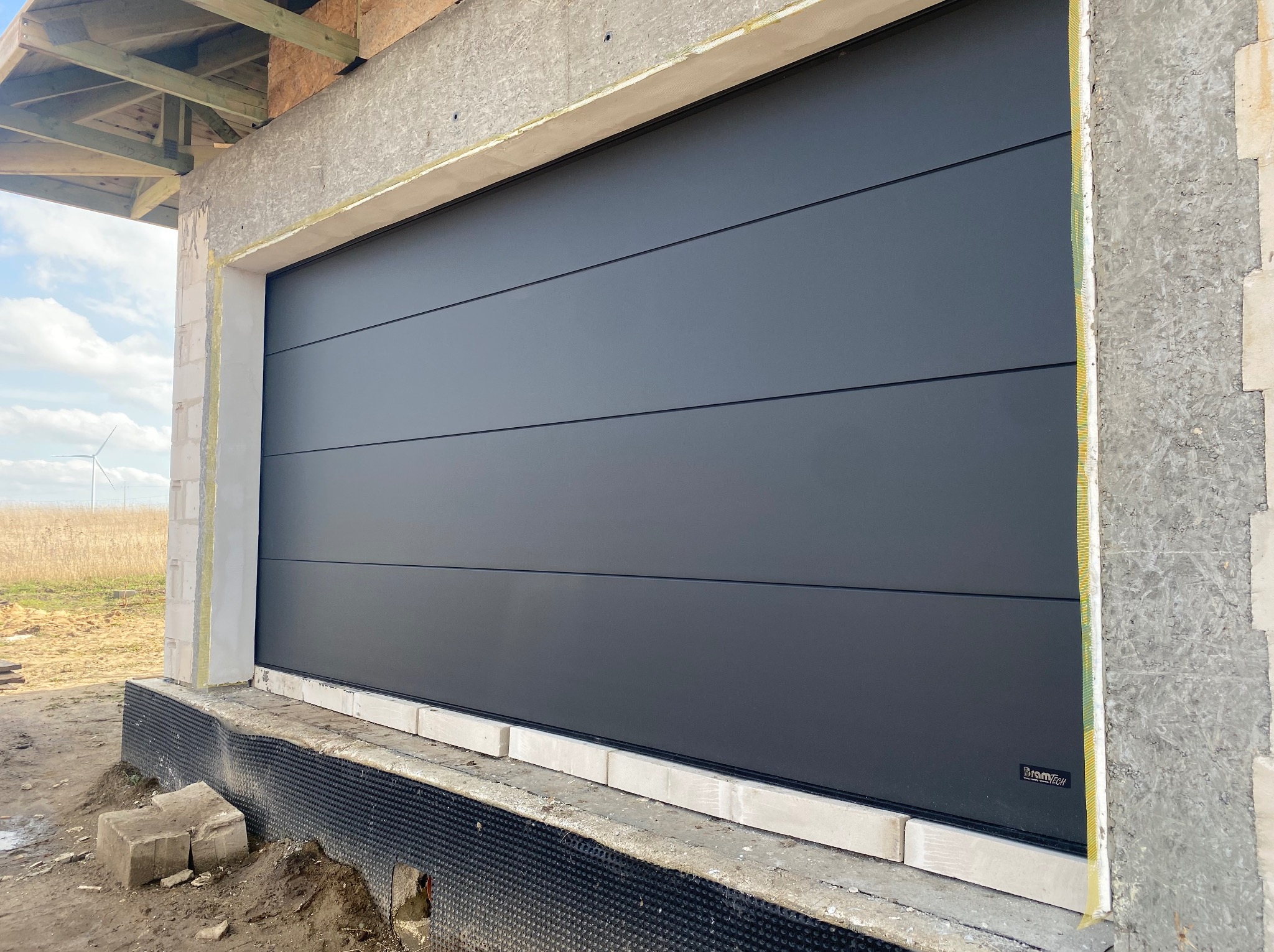 Szara segmentowa brama garażowa z poziomymi przetłoczeniami, zamontowana w surowym otworze budynku w trakcie budowy, widoczne bloczki betonowe i izolacja, w tle pole i turbina wiatrowa.