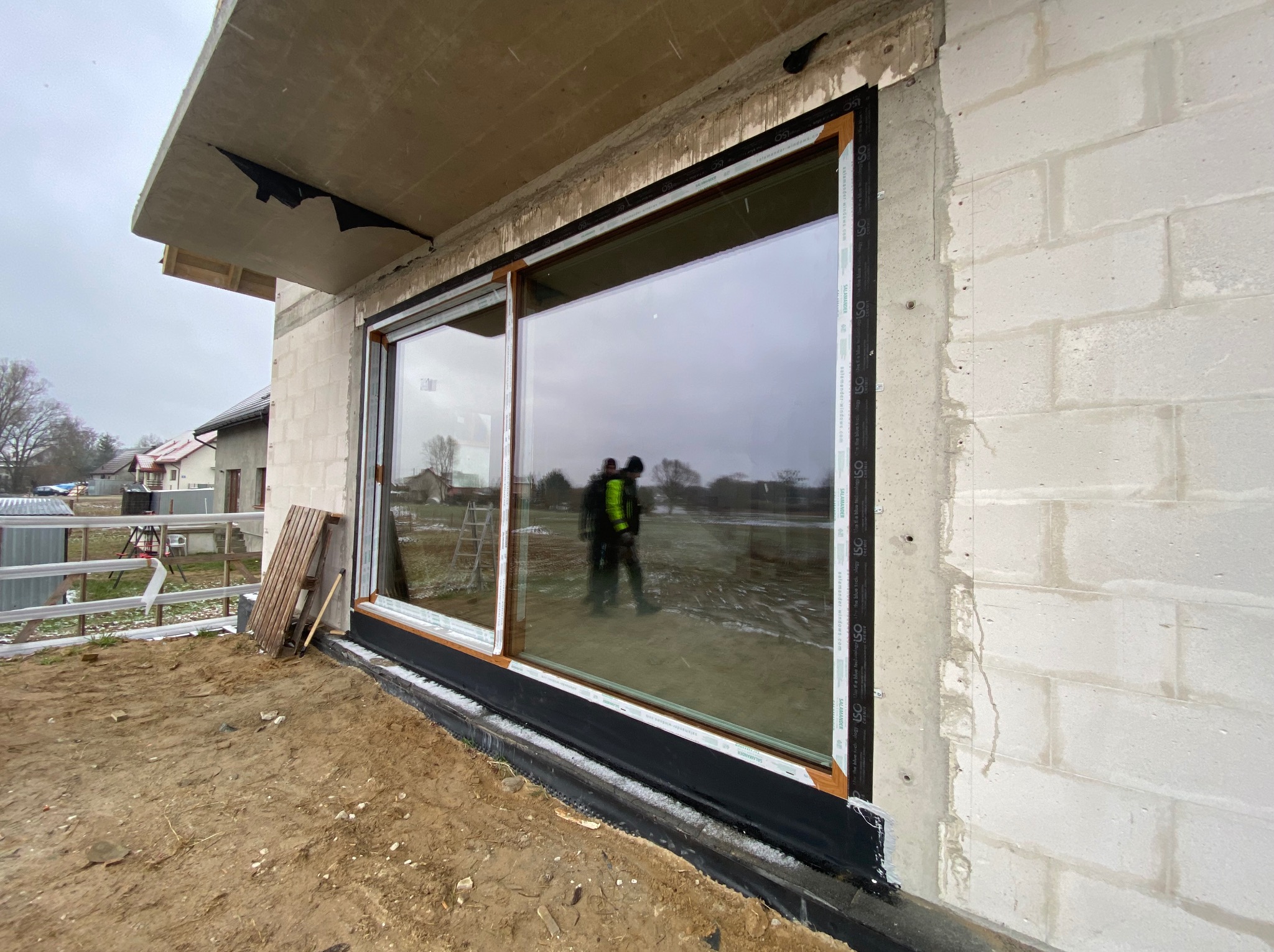 Montaż dużego okna przesuwnego z brązową ramą w surowym stanie budynku, widoczne narzędzia i odbicie dwóch osób w szybie.