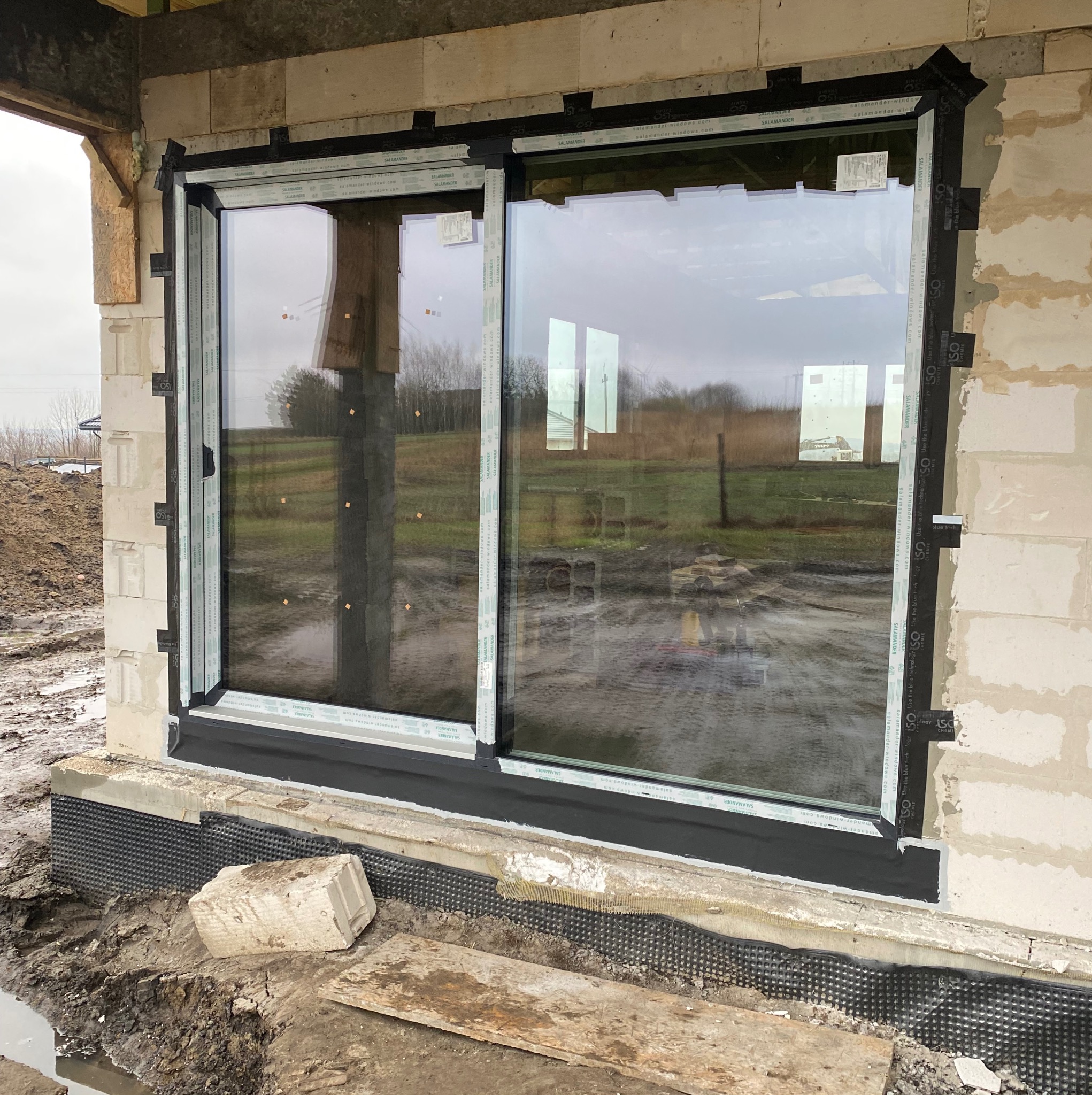 Montaż okna przesuwnego z czarną ramą w surowym stanie budynku, widoczne bloczki betonowe i folia hydroizolacyjna, na szybie odbija się krajobraz.