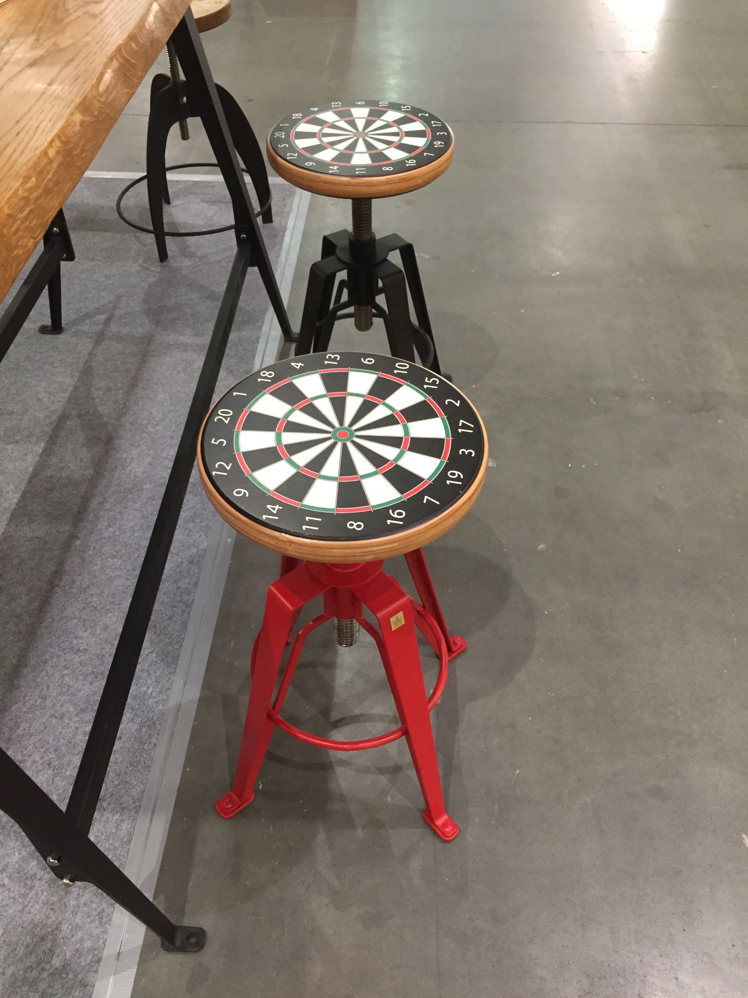 Dwa regulowane stołki barowe z siedziskami w kształcie tarczy do darta, jeden z czarną ramą, drugi z czerwoną, stojące obok drewnianego blatu stołu z metalową nogą w industrialnym stylu.