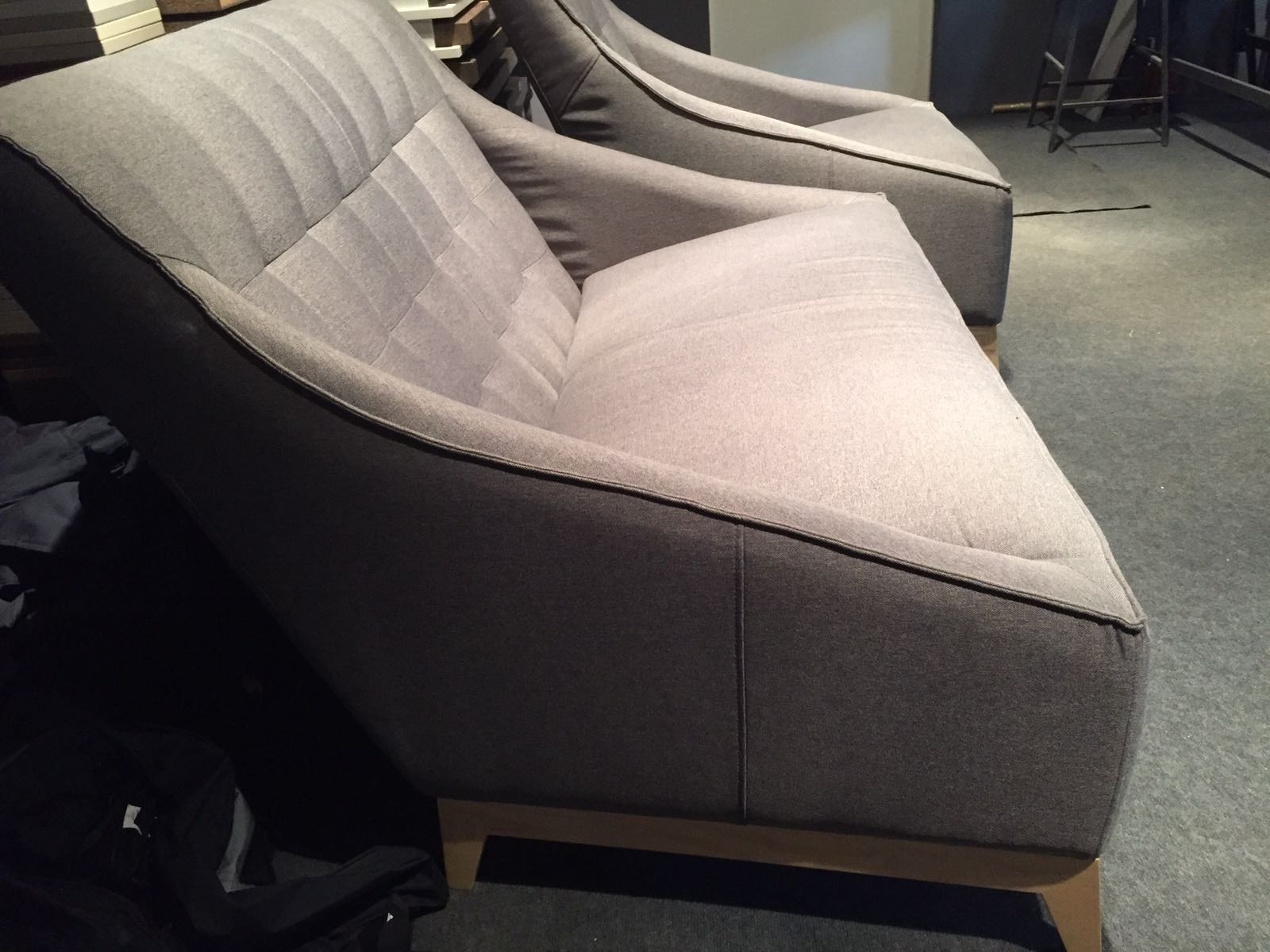 Dwa szare, tapicerowane fotele z niskim oparciem i drewnianymi nóżkami, ustawione jeden za drugim, z widocznym pikowaniem na oparciu jednego z foteli.