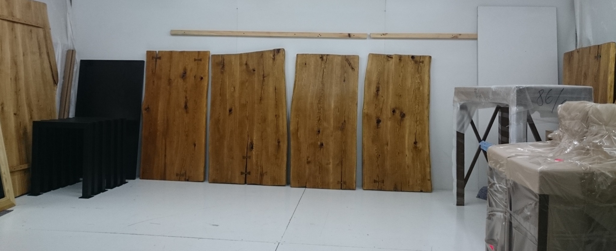 Surowe, drewniane blaty z widocznymi sękami i wstawkami motylkowymi leżące na podłodze, obok czarne, minimalistyczne nogi do stołu i zapakowane stoły z metalowymi nogami w tle.